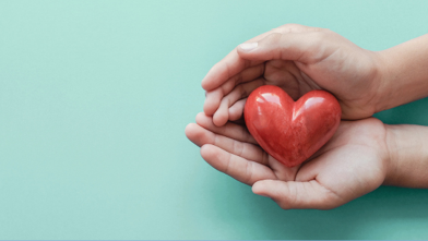 Hands gently cradle a shiny red heart on a light teal background.