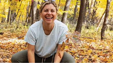 A woman in a gray t-shirt crouches on autumn leaves in a forest, smiling joyfully. Surrounding trees have vibrant yellow foliage.