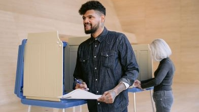 Smiling bearded man exits a voting booth holding a ballot, while a woman votes behind him in a bright polling station.