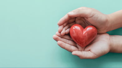 Hands gently cradle a shiny red heart on a light teal background.