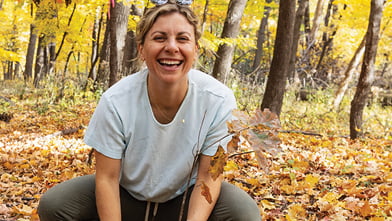 A woman in a gray t-shirt crouches on autumn leaves in a forest, smiling joyfully. Surrounding trees have vibrant yellow foliage.