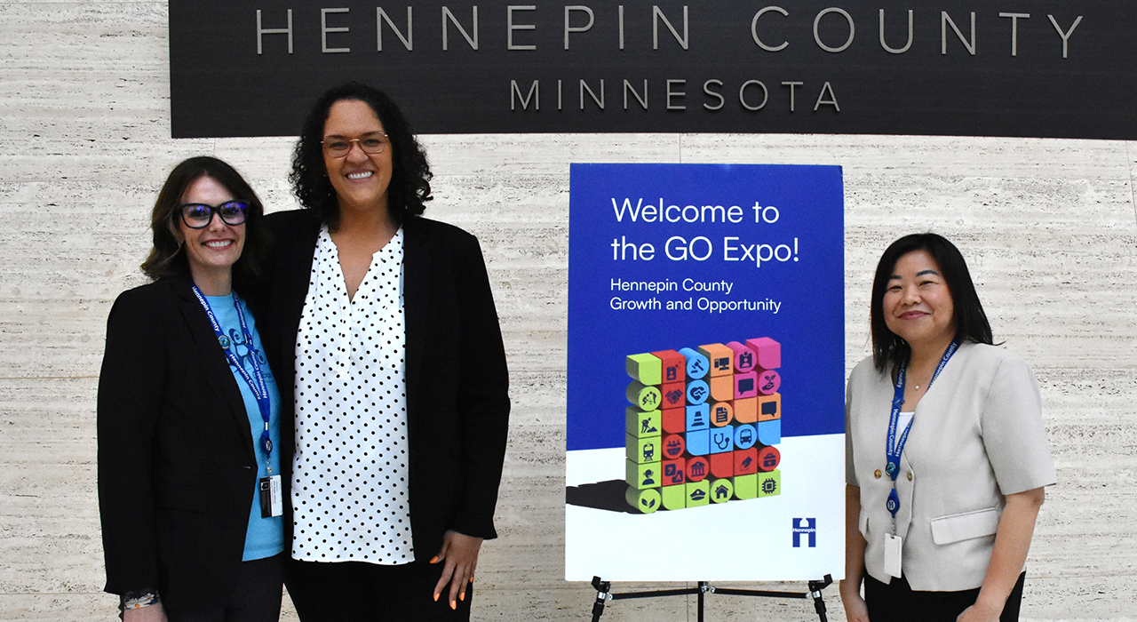 Three people stand beside a welcome sign for the GO Expo inside a Hennepin County building.