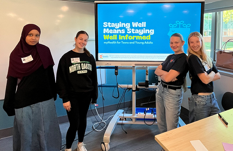 Four students in a classroom standing in front of a screen that says "Staying Well Means Staying Well Informed".